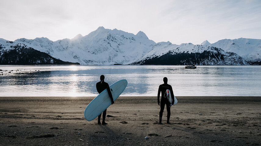 Zwei Männer im Neoprenanzug und Surfboards stehen an einem Strand und schauen auf schneebedeckte Berge