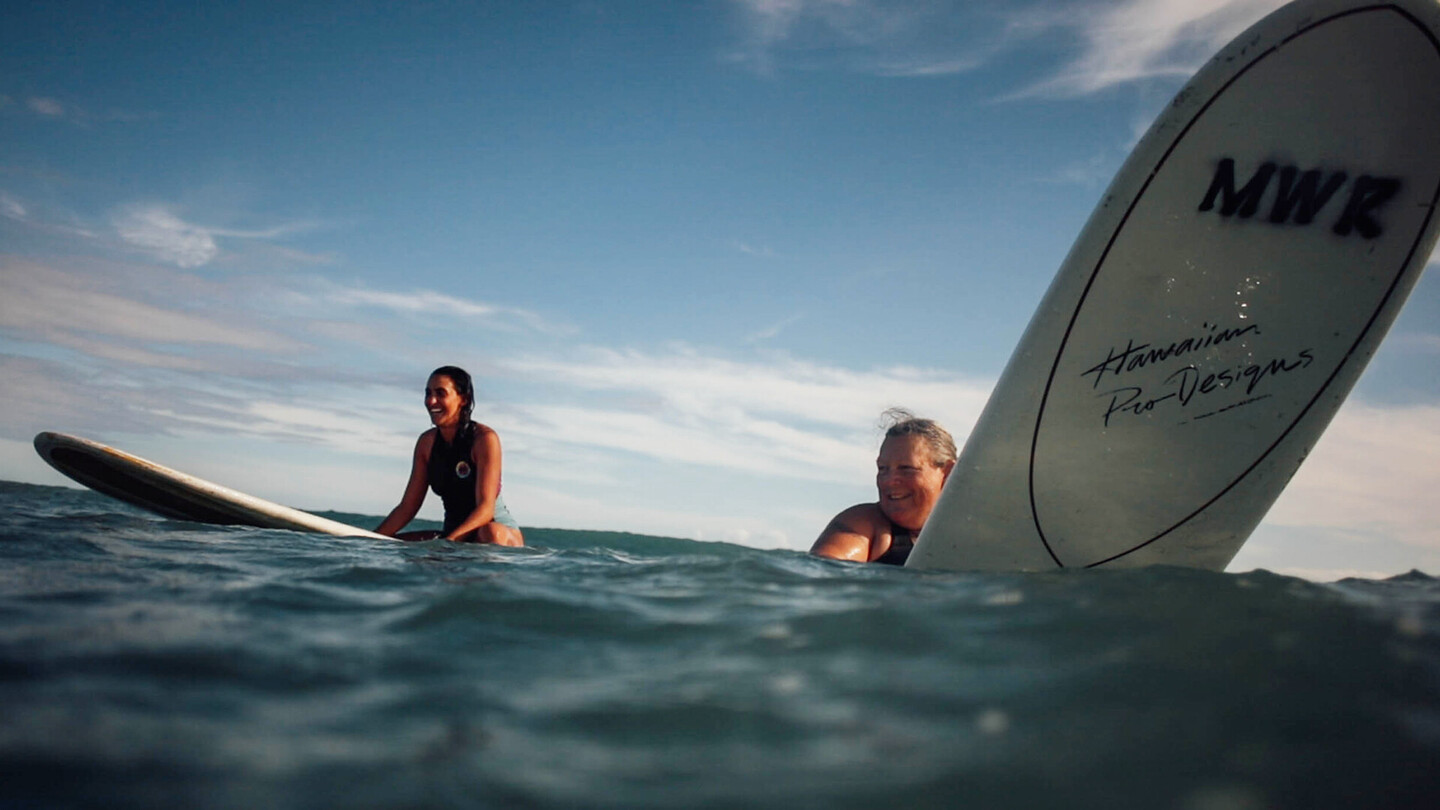 Zwei lachende Surferinnen sitzen auf ihren Boards im offenen Meer, bereit für die nächste Welle.