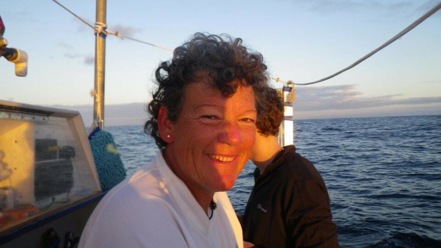 Frau mit kurzen Locken und weißem Shirt sitzt auf einem Boot bei Sonnenuntergang, mit Blick auf das offene Meer.